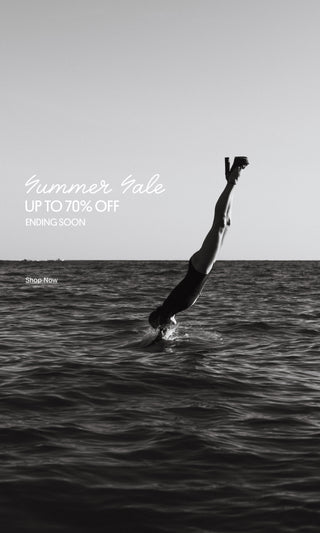 Person diving headfirst into the ocean while wearing black platform heels, with a monochrome seascape in the background.