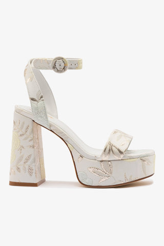 An ivory brocade sandal with an open round toe, adjustable ankle strap, and a high block heel.